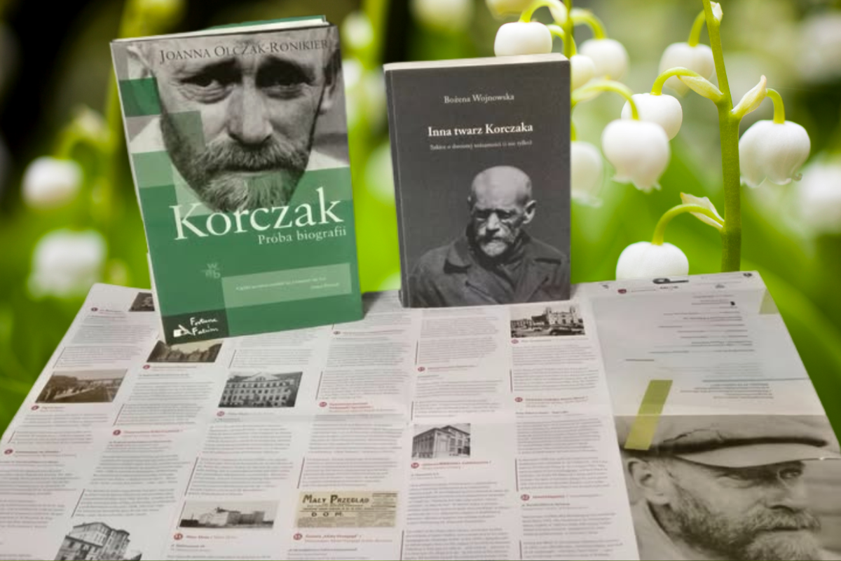 Dwie książki "Korczak" Joanny Olczak-Ronikier i "Inna twarz Korczaka" Bożeny Wojciszke leżą na stole z rozłożonym informacyjnym folderem, w tle zielone liście i białe kwiaty.